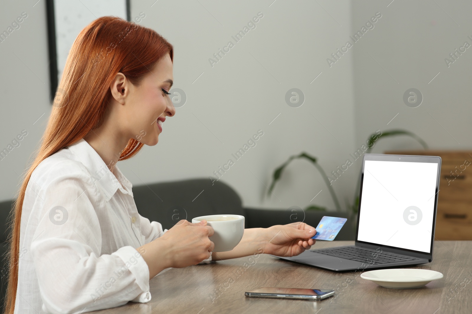 Photo of Happy woman with credit card and cup of drink near laptop in room. Online shopping