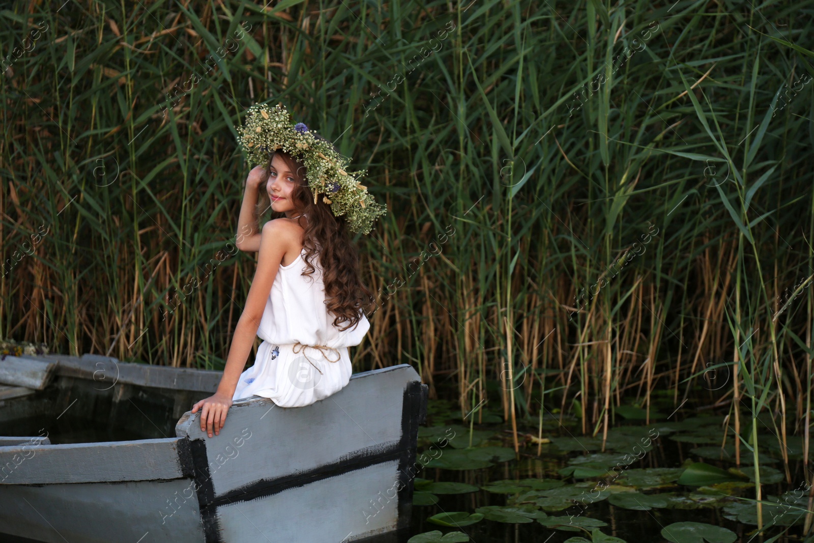 Photo of Cute little girl wearing wreath made of beautiful flowers in wooden boat