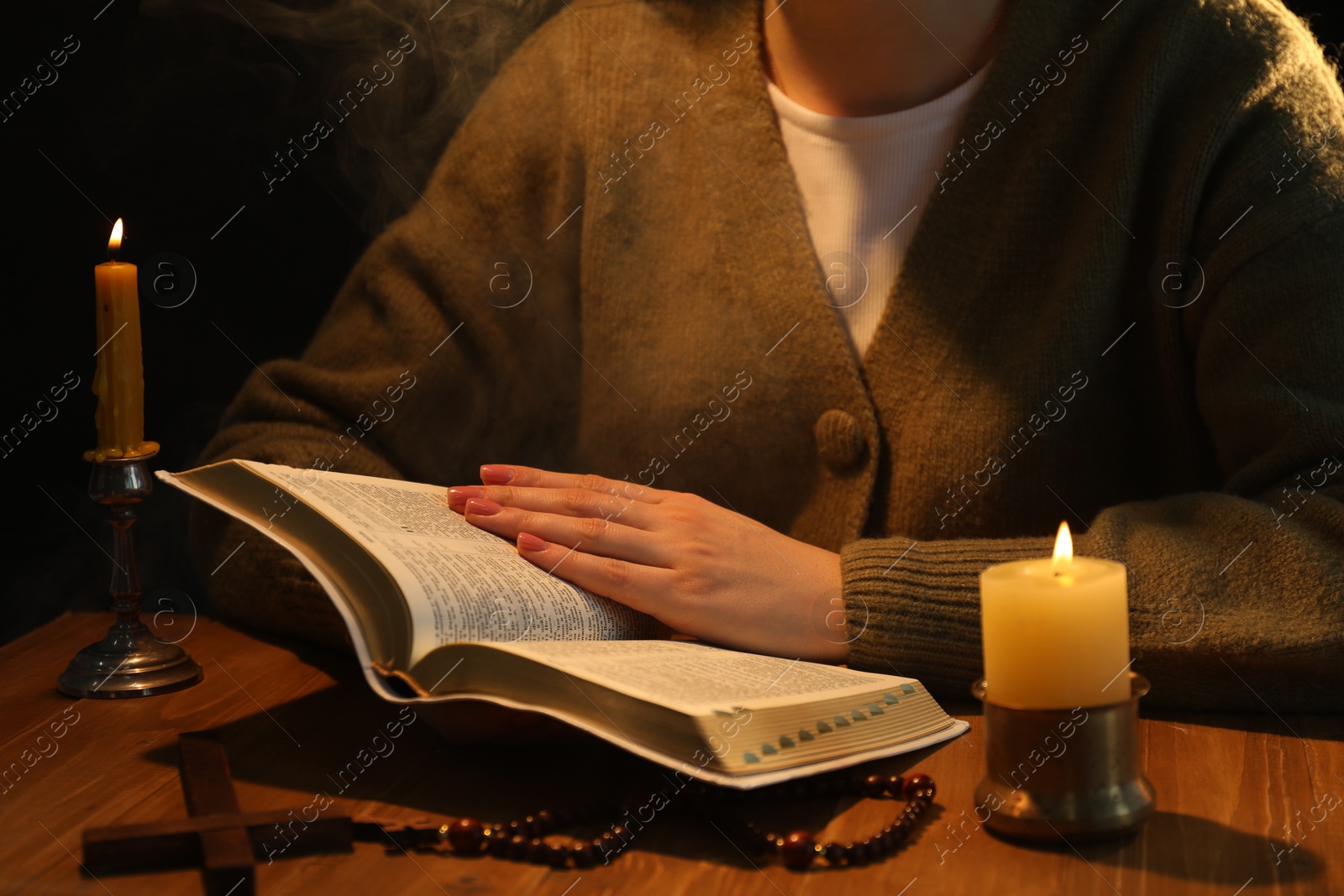 Photo of Woman reading Bible at table with burning candles, closeup