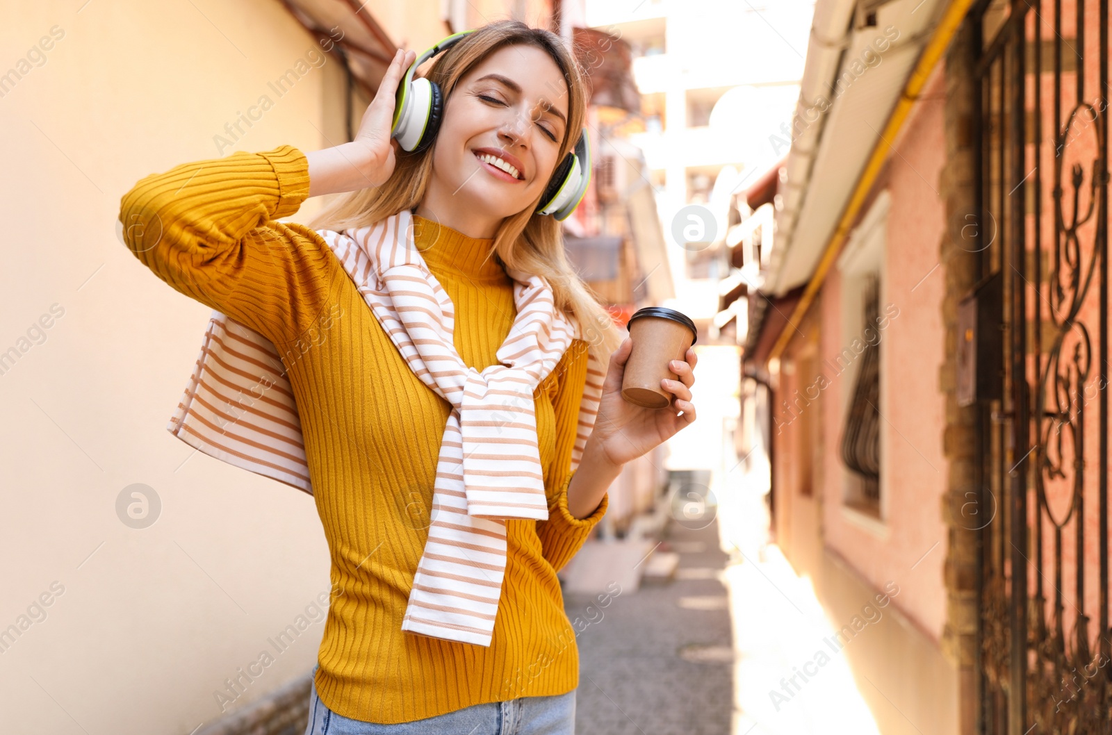 Photo of Happy young woman with coffee and headphones listening to music on city street