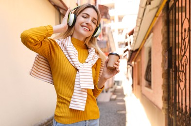 Photo of Happy young woman with coffee and headphones listening to music on city street