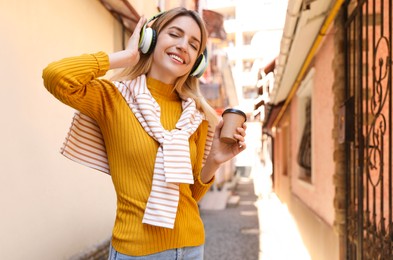 Happy young woman with coffee and headphones listening to music on city street