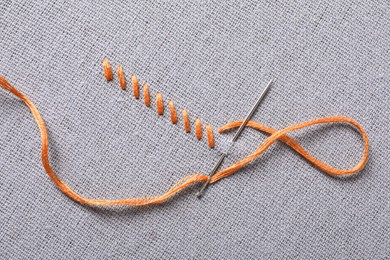 Photo of Needle with orange embroidery floss and row of stitches on grey fabric, top view
