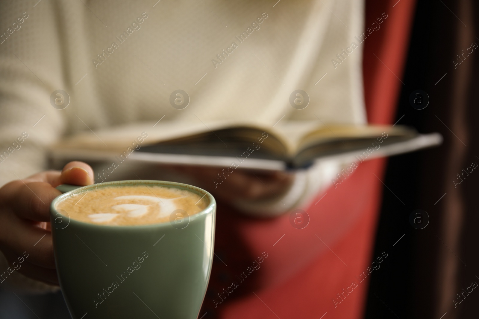 Photo of Woman with coffee reading book indoors, focus on cup