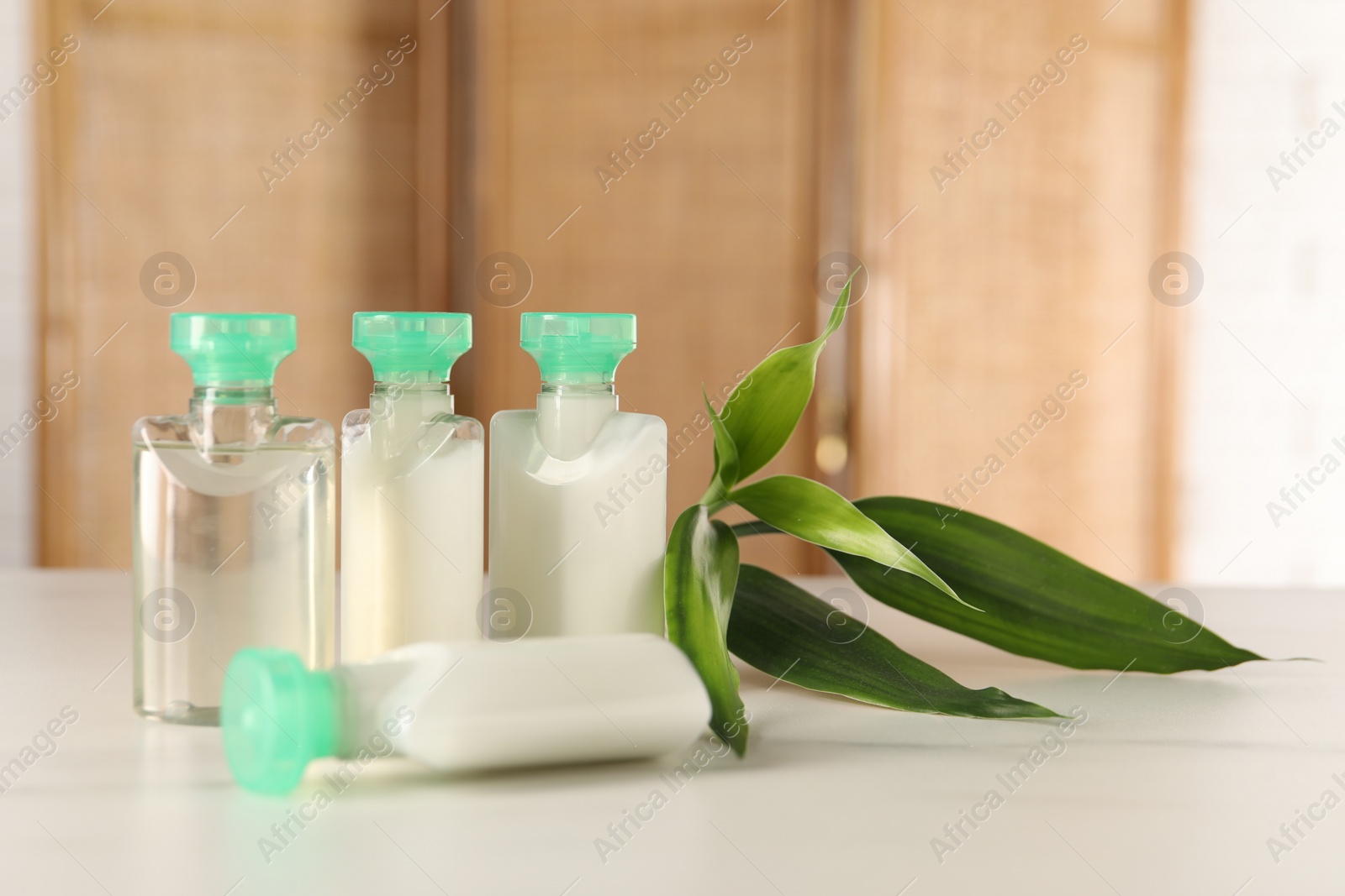 Photo of Mini bottles of cosmetic products and green branch on white table against blurred background. Space for text