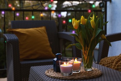 Soft pillows, blanket, burning candles and yellow tulips on rattan garden furniture in evening