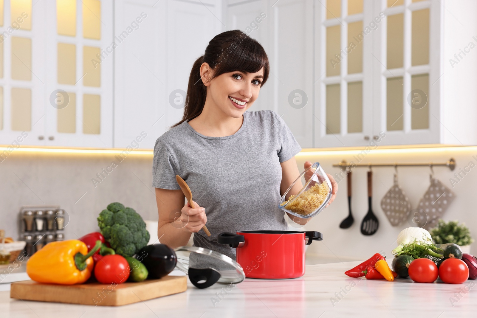 Photo of Happy young housewife adding raw pasta in pot at white marble table in kitchen. Cooking process