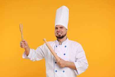 Photo of Happy professional confectioner in uniform holding rolling pin and spatula on yellow background