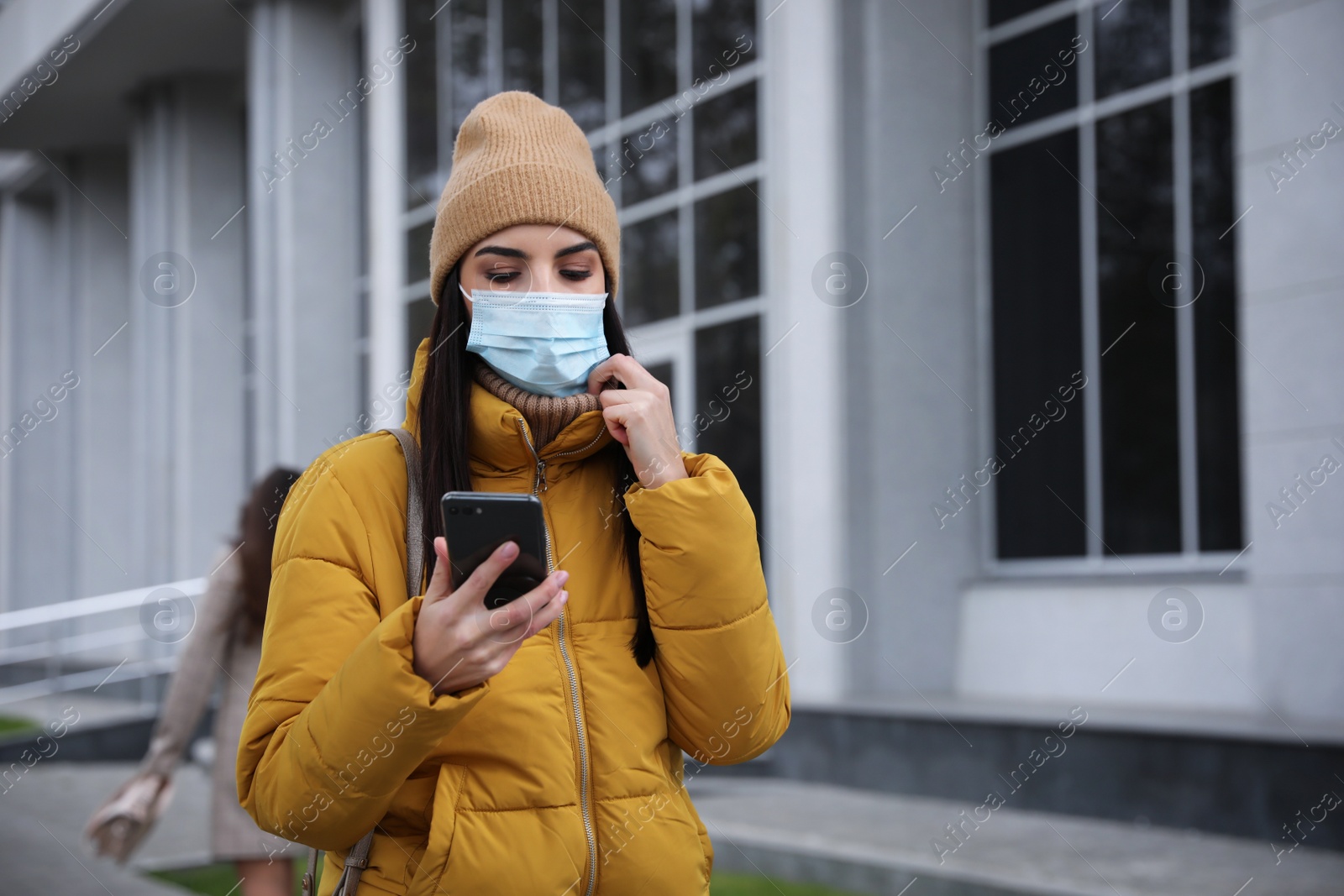 Photo of Young woman in medical face mask with smartphone walking outdoors. Personal protection during COVID-19 pandemic