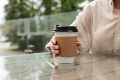 Photo of Woman holding paper takeaway cup at glass table outdoors, closeup. Coffee to go