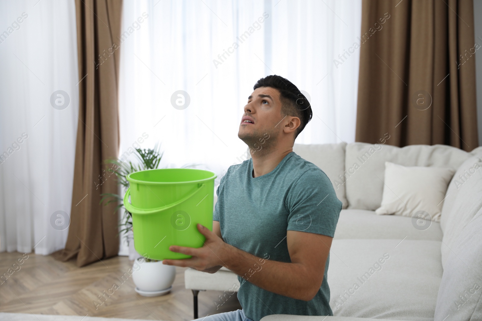 Photo of Emotional man collecting water leaking from ceiling in living room. Damaged roof