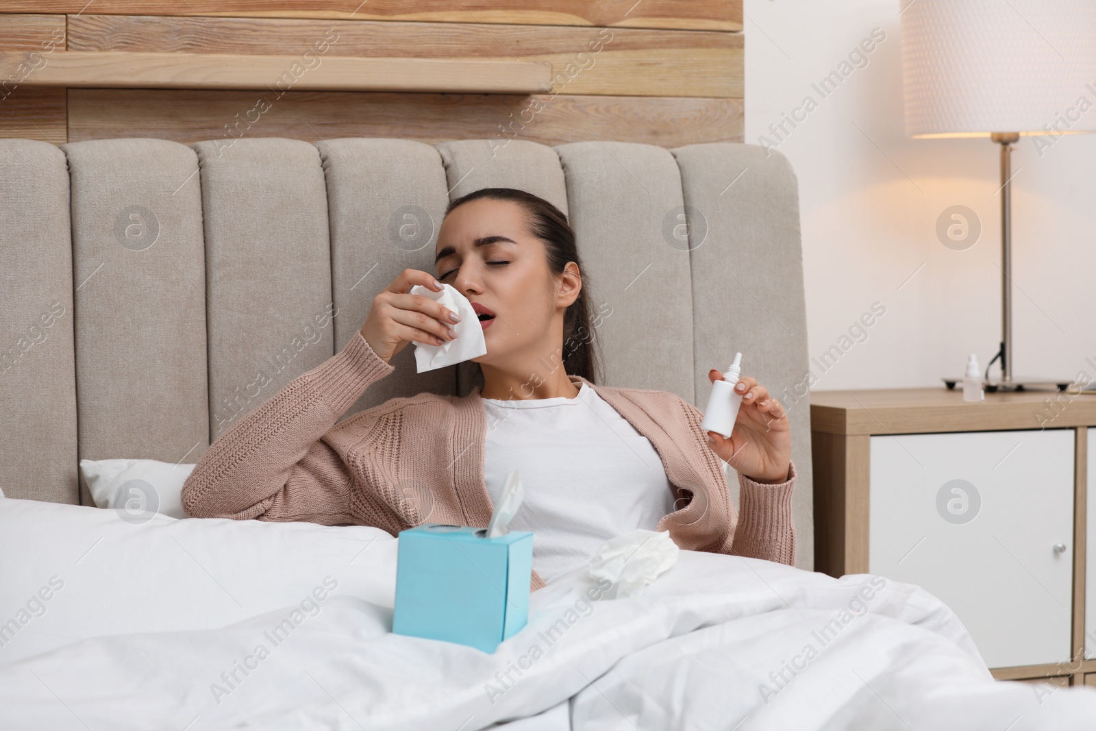 Photo of Sick young woman with napkins and nasal spray in bed at home