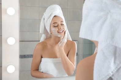 Photo of Young woman washing face with soap near mirror in bathroom