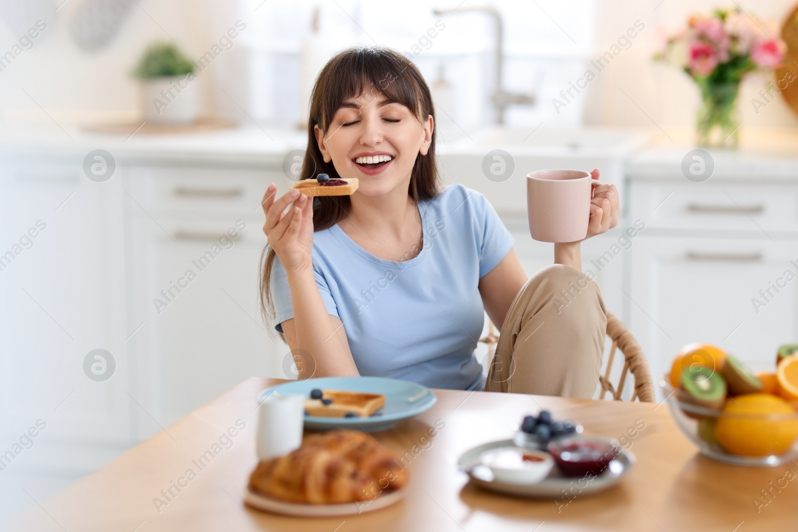 Photo of Smiling woman drinking coffee and eating toast at breakfast indoors