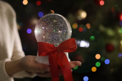 Woman holding snow globe with red bow knot on blurred background, closeup. Space for text