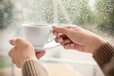 Photo of Young woman with cup of coffee near window indoors on rainy day, closeup