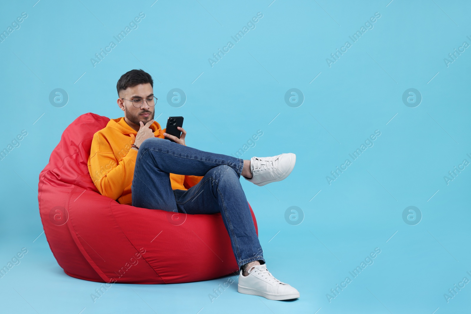 Photo of Handsome young man using smartphone on bean bag chair against light blue background. Space for text