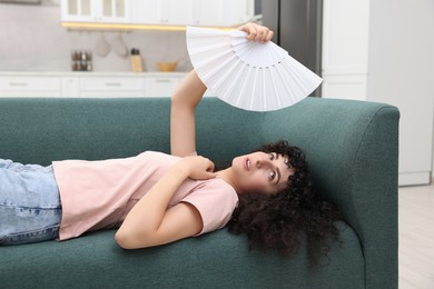 Photo of Young woman waving white hand fan to cool herself on sofa at home