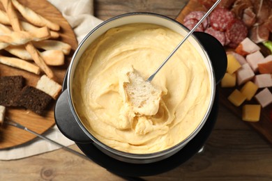 Dipping piece of bread into fondue pot with melted cheese at wooden table with snacks, flat lay