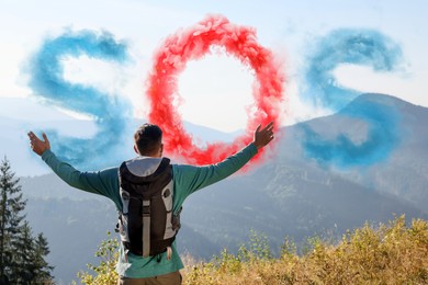 Man with backpack and word SOS made of color smoke bomb in mountains, back view