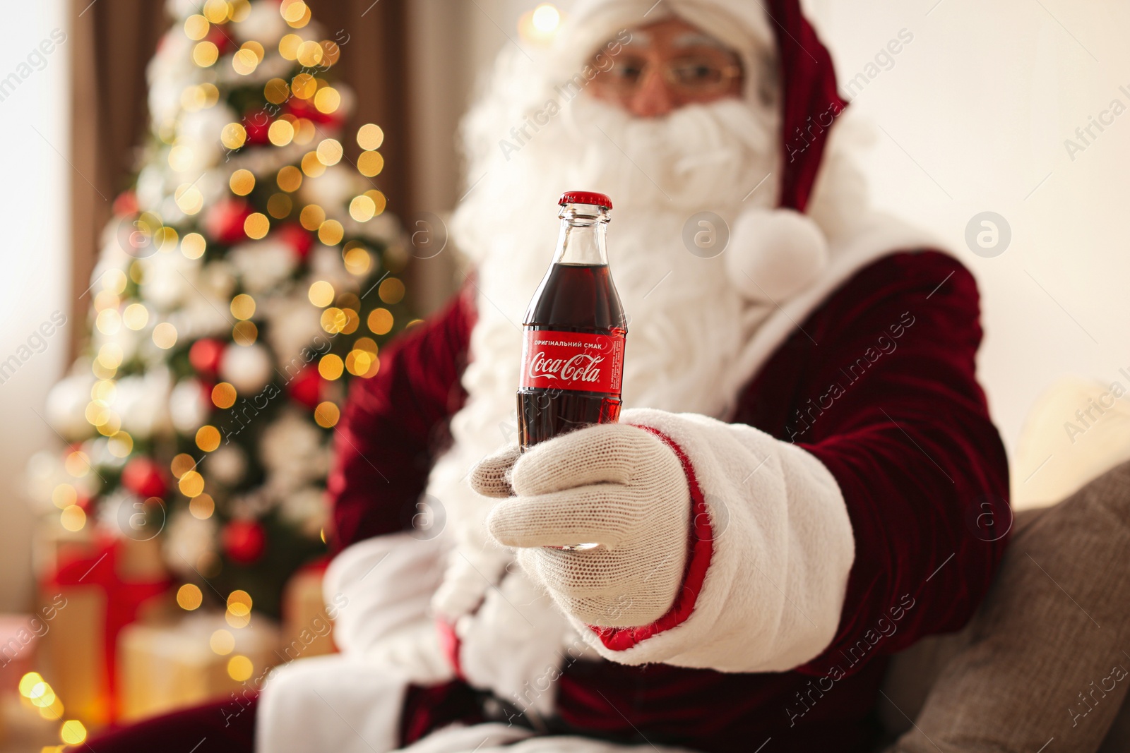 Photo of MYKOLAIV, UKRAINE - JANUARY 18, 2021: Santa Claus with Coca-Cola bottle in room decorated for Christmas, focus on hand