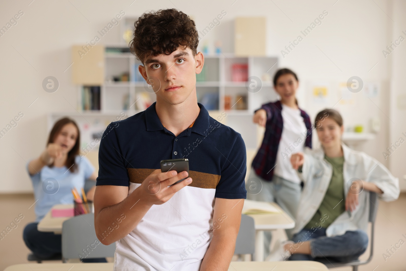 Photo of Teen problems. Lonely boy with smartphone standing separately from other students in classroom