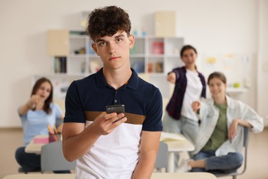 Teen problems. Lonely boy with smartphone standing separately from other students in classroom