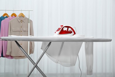 Photo of Modern iron and white shirt on board near rack with clean clothes indoors