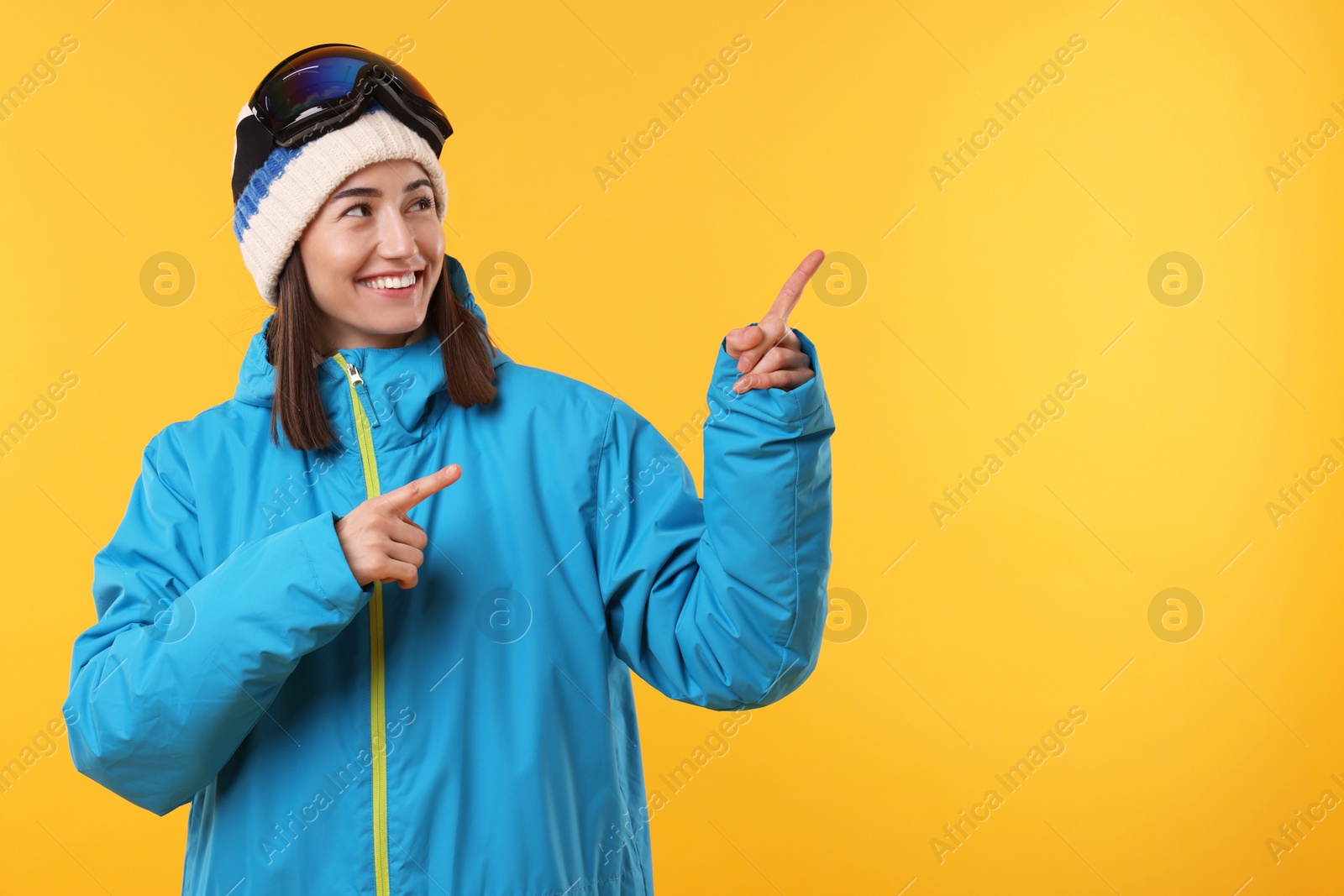 Photo of Winter sports. Happy woman with snowboard goggles pointing at something on orange background, space for text