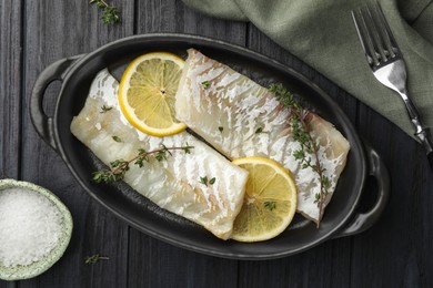 Photo of Fresh raw cod fillets with thyme and lemon in baking dish on black wooden table, flat lay
