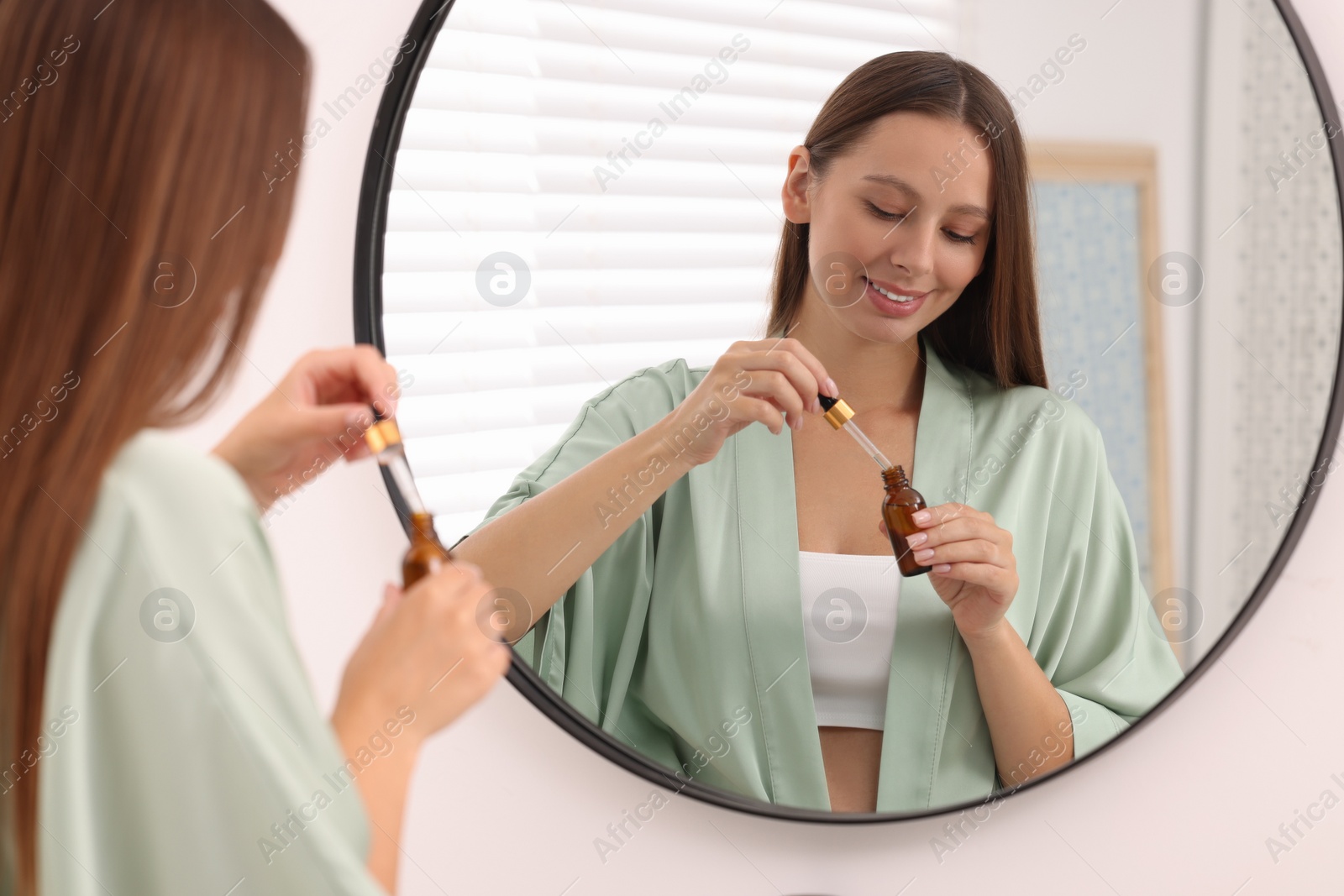 Photo of Beautiful woman holding bottle of serum and dropper near mirror indoors