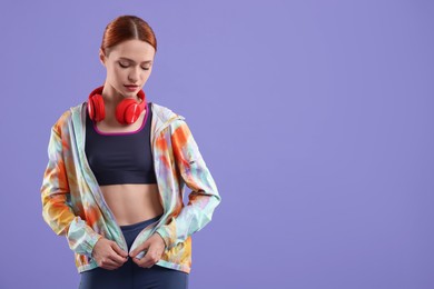 Young woman in sportswear and headphones on violet background, space for text