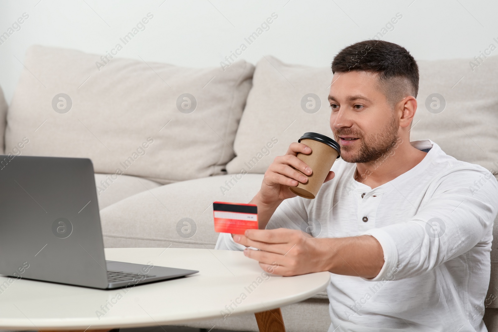 Photo of Happy man with credit card and cup of coffee near laptop at white table indoors. Online shopping