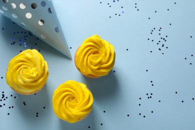 Delicious cupcakes with bright cream, party hat and confetti on light blue background, flat lay. Space for text