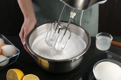 Woman making whipped cream with hand mixer at black table, closeup