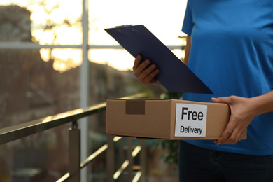 Photo of Courier holding parcel with sticker "Free Delivery"  and clipboard indoors, closeup