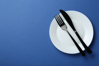 Clean white plate with cutlery on blue background, top view. Space for text