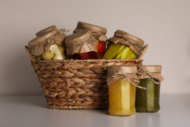 Photo of Wicker basket with many jars of different preserved products on light grey table