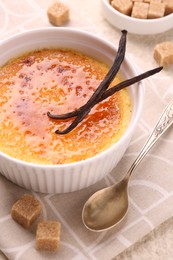 Photo of Delicious creme brulee in bowl, vanilla pods, sugar cubes and spoon on table, closeup