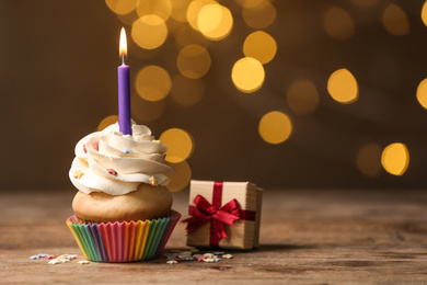 Photo of Birthday cupcake with candle and gift box on wooden table against blurred lights. Space for text