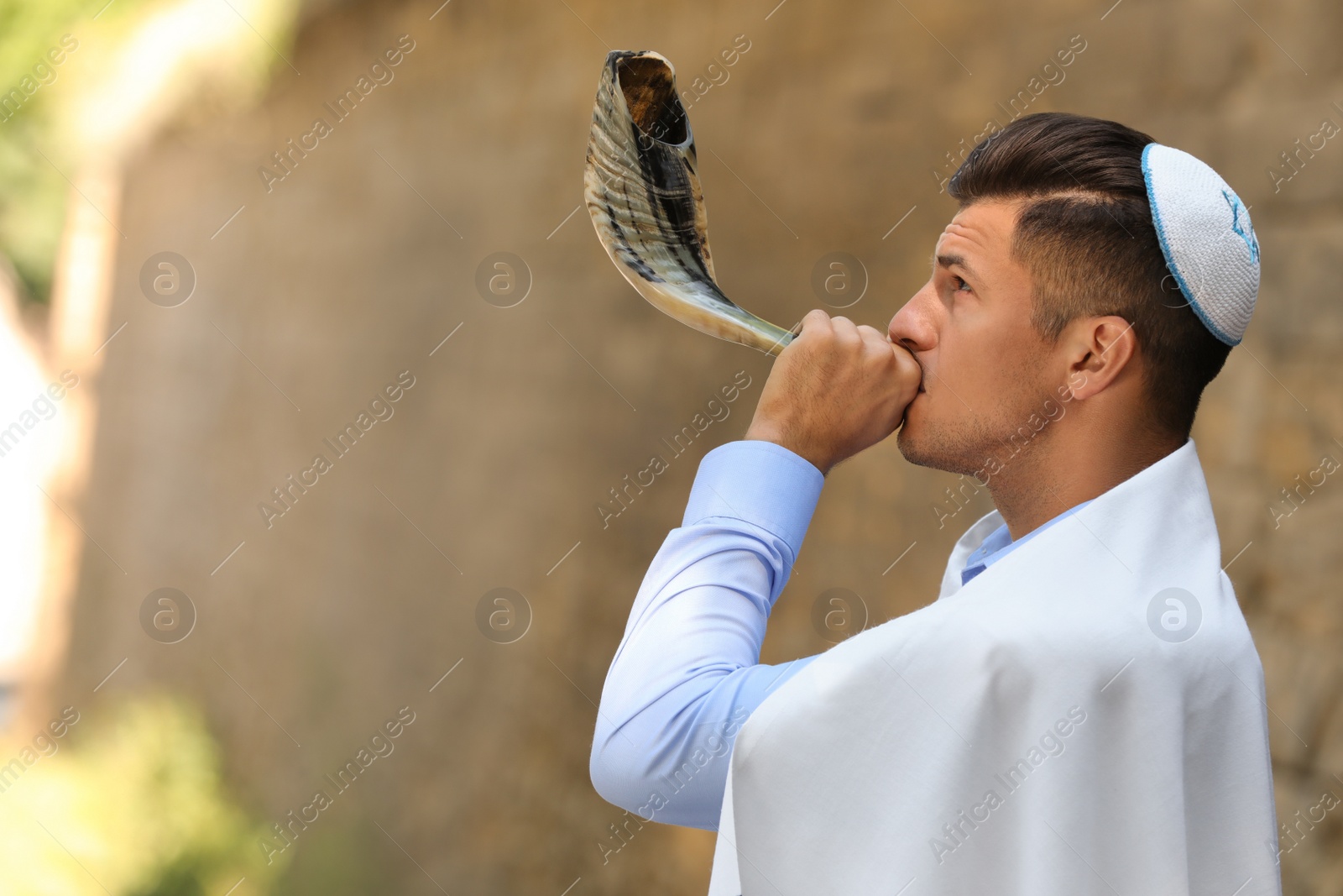 Photo of Jewish man in kippah and tallit blowing shofar outdoors. Rosh Hashanah celebration