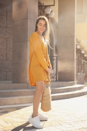 Beautiful young woman in stylish yellow dress with handbag on city street