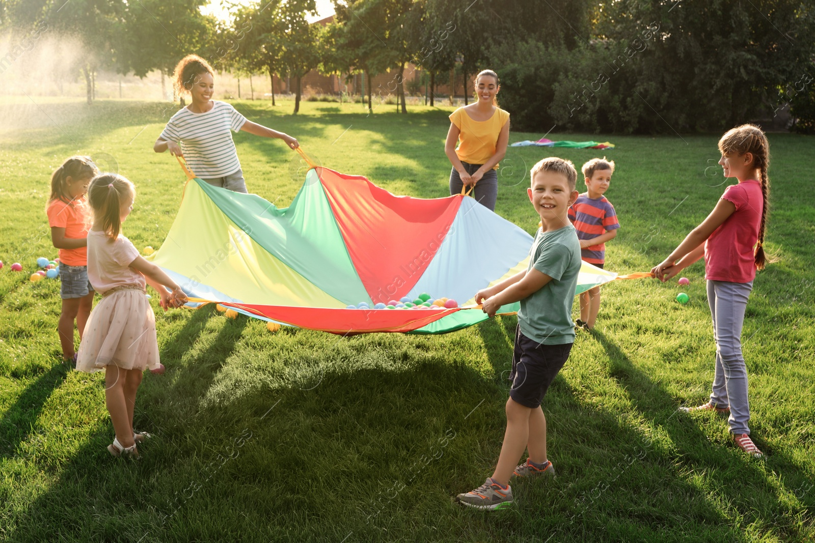 Photo of Group of children and teachers playing with rainbow playground parachute on green grass. Summer camp activity