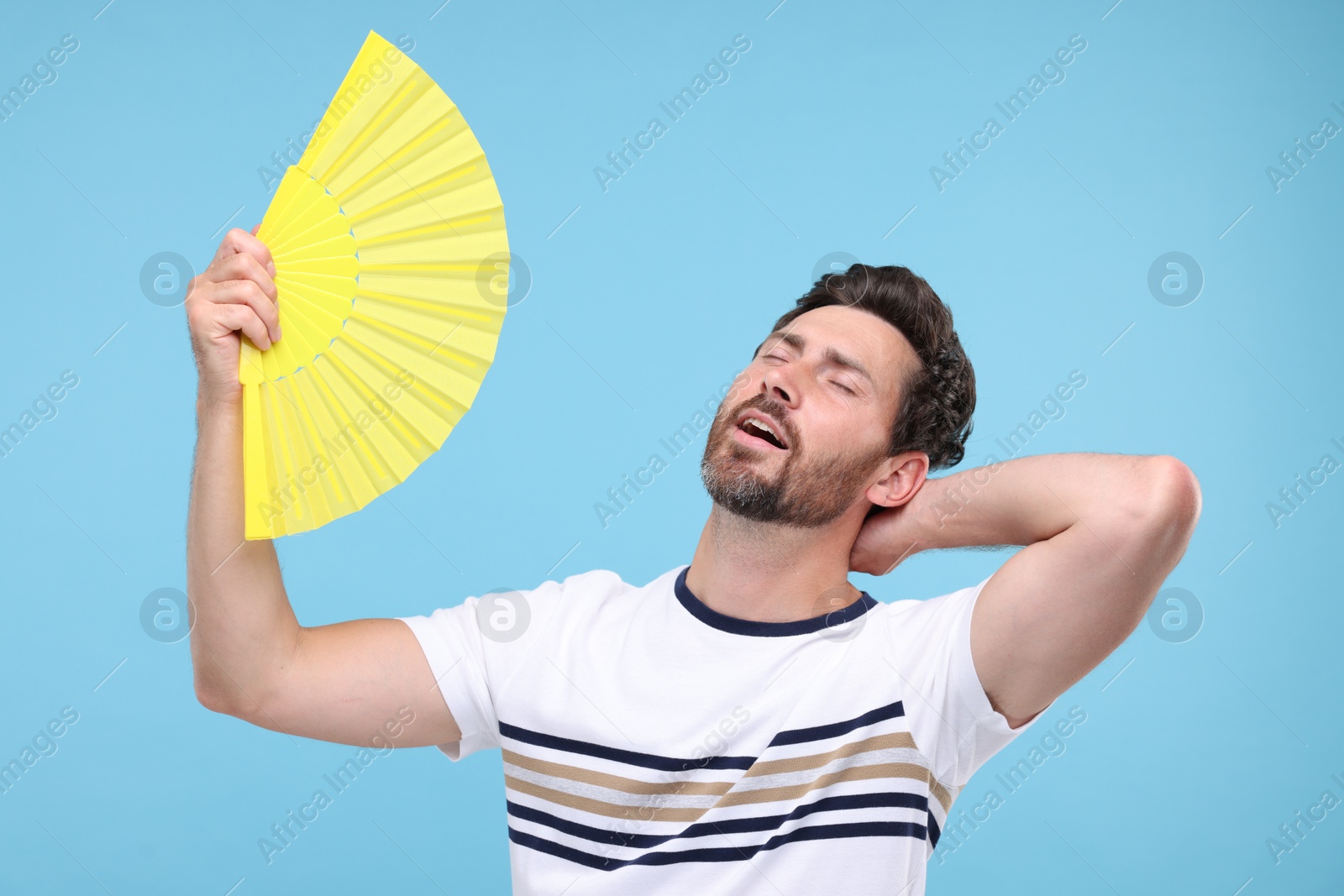 Photo of Unhappy man with hand fan suffering from heat on light blue background