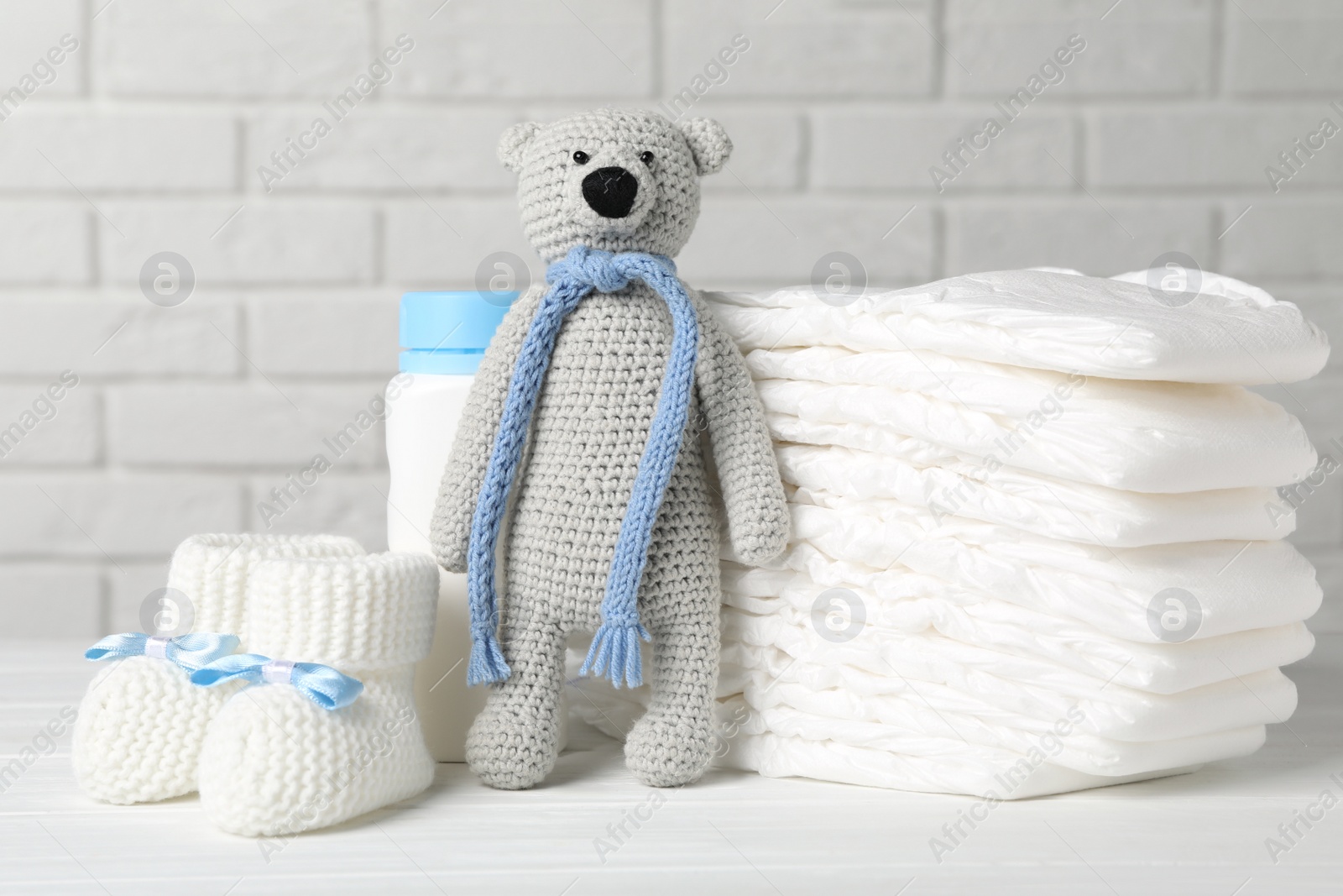 Photo of Baby diapers, toy bear, booties and bottle on wooden table against white brick wall