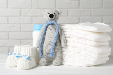 Photo of Baby diapers, toy bear, booties and bottle on wooden table against white brick wall