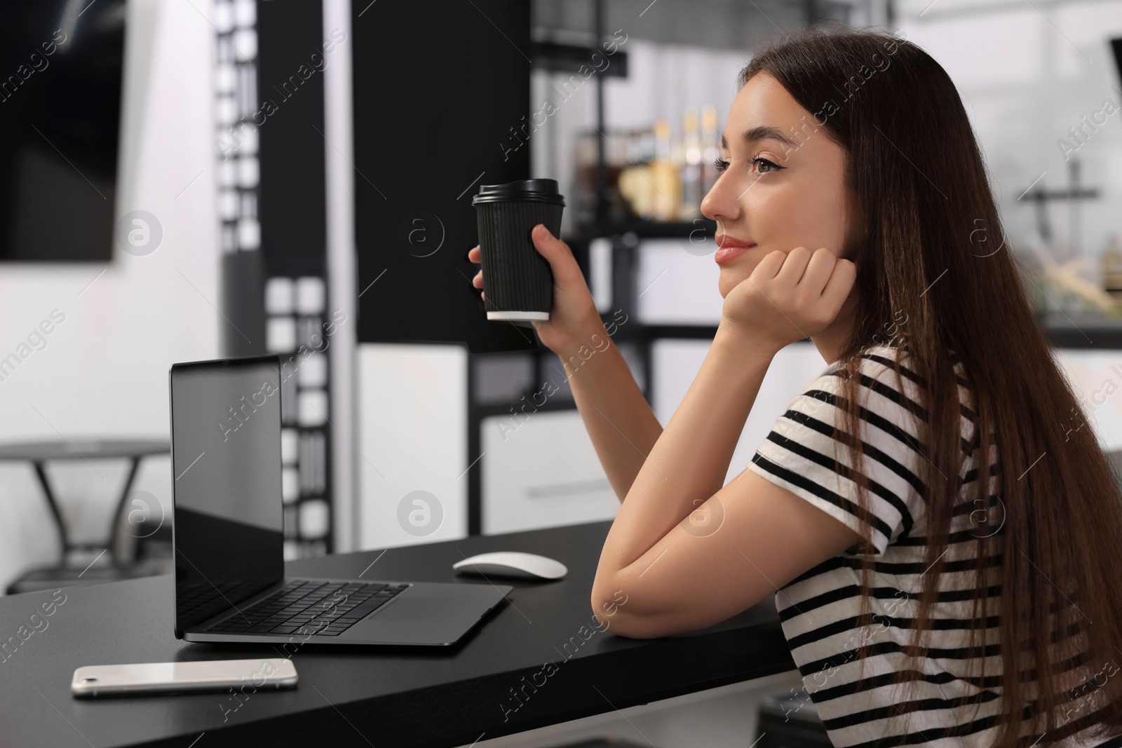 Photo of Young woman with paper cup of coffee and laptop at table in hostel dining room