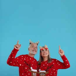 Photo of Senior couple in Christmas sweaters, reindeer headband and party glasses pointing at something on light blue background