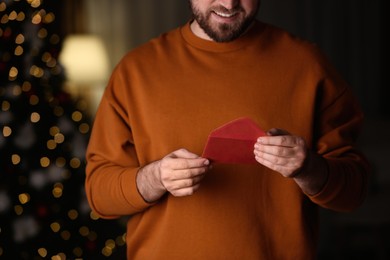 Photo of Man holding envelope with greeting card against blurred Christmas lights, closeup