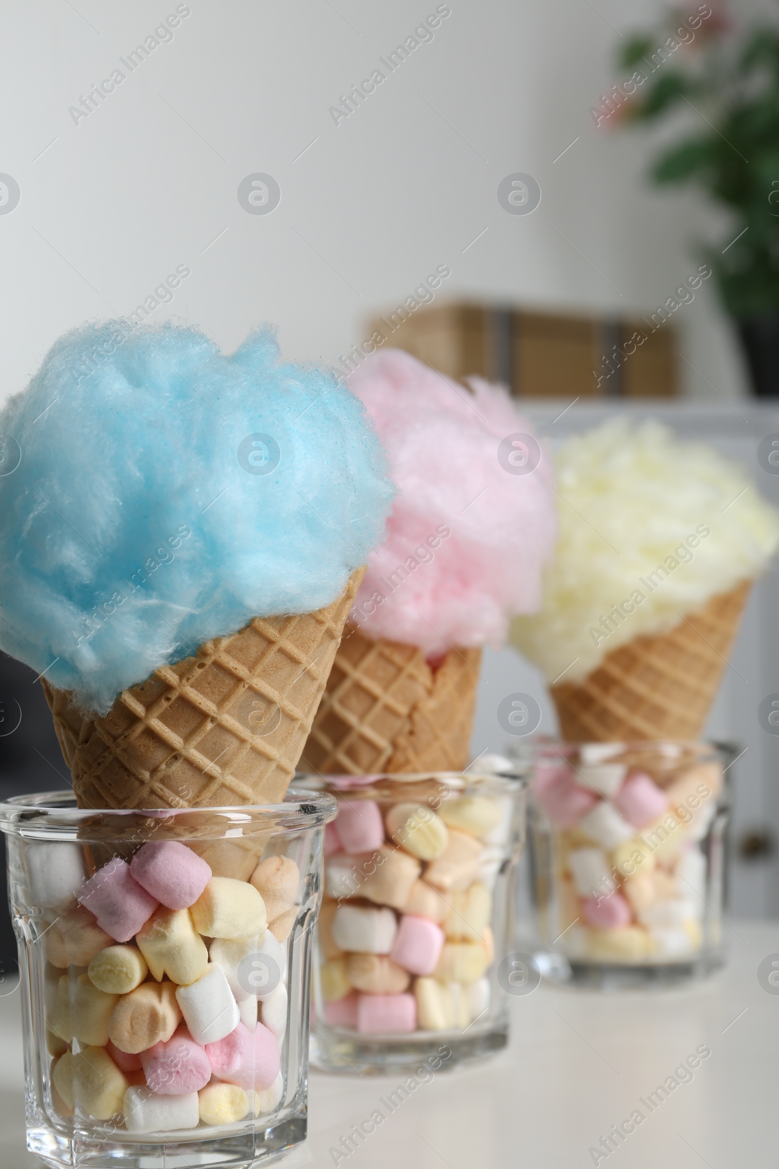 Photo of Sweet cotton candies in waffle cones and marshmallows on white table indoors, closeup